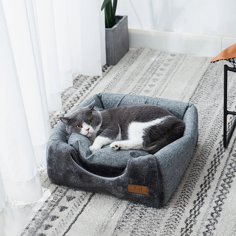 Cat lying on a gray pet bed in a room with white curtains and a plant.