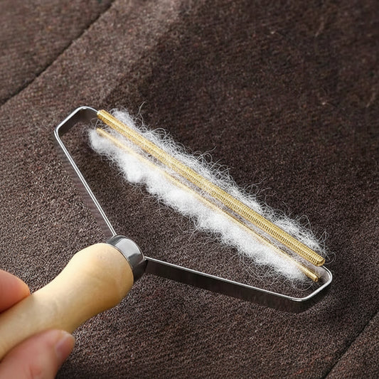 Brass coil fabric shaver lifting white pet hair off a brown wool coat, with collected hair piled on the fabric beside it