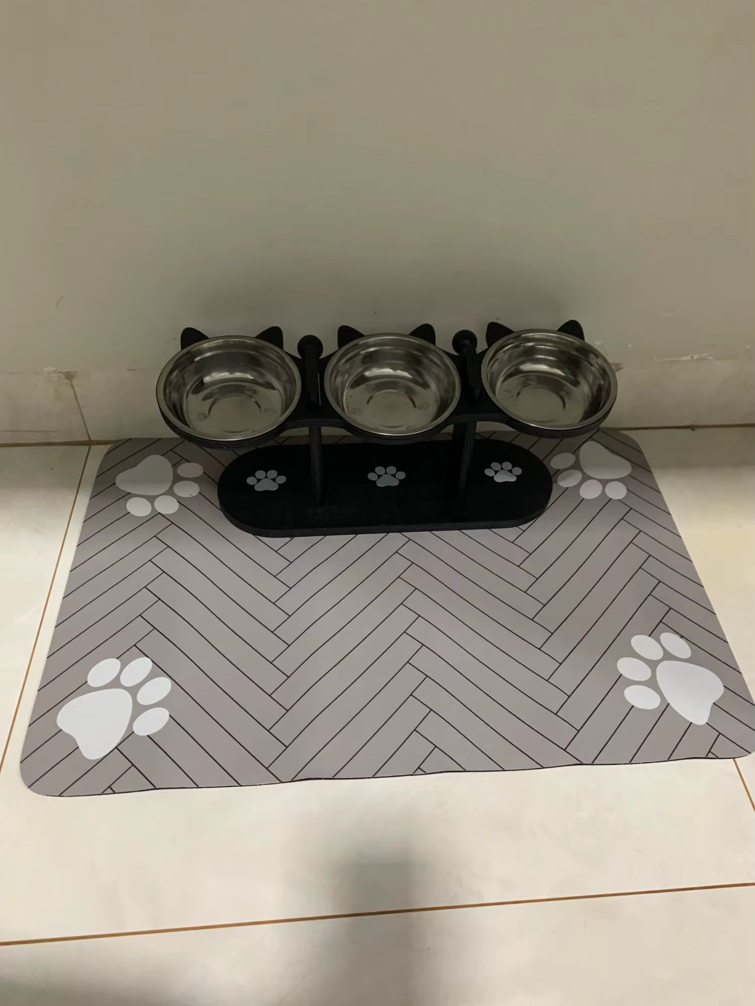 Pet feeding station with three bowls on a mat with paw prints on a tiled floor.
