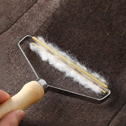 Brass coil fabric shaver lifting white pet hair off a brown wool coat, with collected hair piled on the fabric beside it