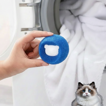 A hand holding a blue PawCatch sponge ball in front of a washing machine drum with a white towel inside.