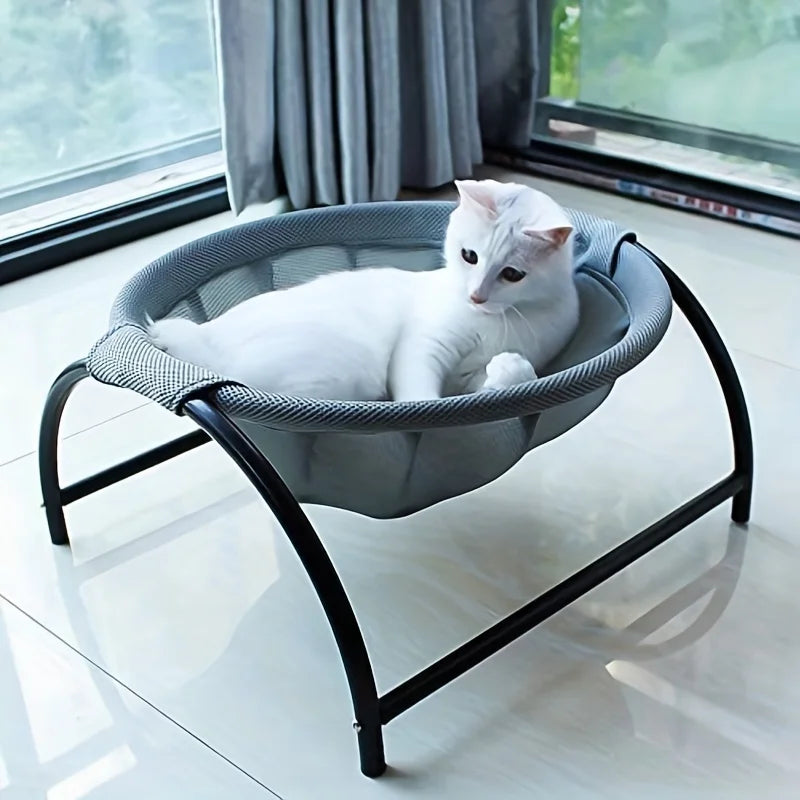 White cat resting in the PawRest grey mesh cat hammock bed on an arched black steel frame, with a plant in the background