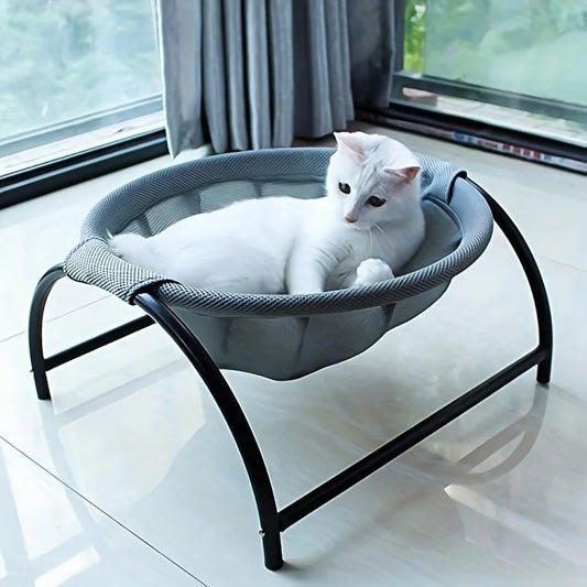 White cat resting in the PawRest grey mesh cat hammock bed on an arched black steel frame, with a plant in the background