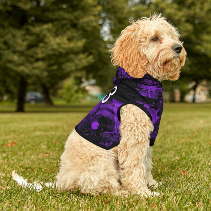 Dog wearing a purple and black patterned coat sitting on grass with trees in the background