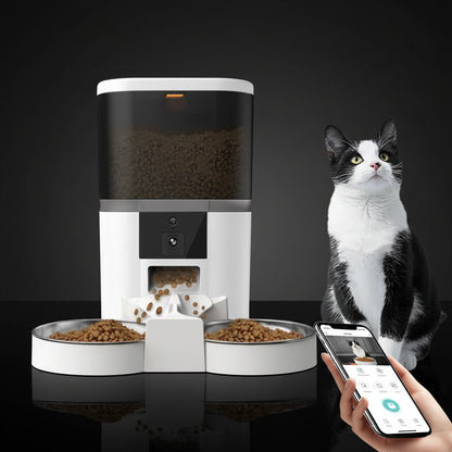 White automatic pet feeder with HD camera and dual stainless steel bowls, beside a smartphone showing the feeder app, black and white cat sat watching. 